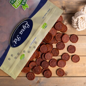 Eine Tüte mit Haustierleckerlis mit der Aufschrift 'Furta Set' kippt runde, rötlich-braune Snacks auf eine Holzoberfläche, mit einem Seilspielzeug in der Nähe.