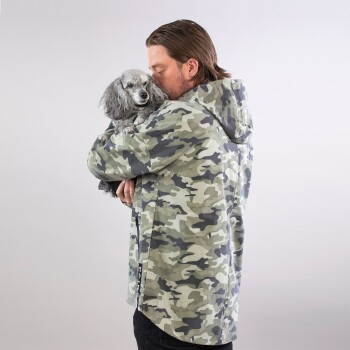 Eine Person in einer Camouflage-Jacke hält einen kleinen Hund nah an ihrer Brust, vor einem schlichten weißen Hintergrund.