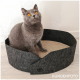 Eine graue Katze mit leuchtend orangefarbenen Augen sitzt in einem runden, dunklen Filz-Katzenbett mit einem strukturierten beigen Boden, vor einer hellen Wand.