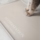 Die Pfoten einer Katze ruhen auf einer beigen Futtermatte mit dem Text 'FUTTERSTATION' und 'kleckern erlaubt' in Weiß gedruckt.