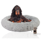 Ein schwarz-brauner Hund, der auf einem flauschigen grauen runden Hundebett ruht, mit einem kleinen beigen Spielzeug neben sich.