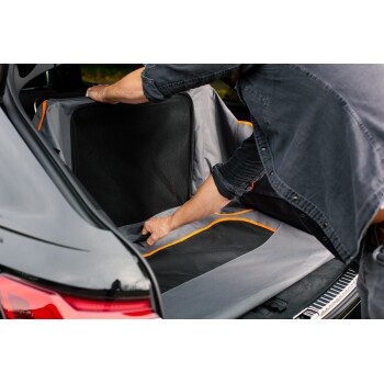 Eine Person legt einen grauen Autositzbezug für Haustiere mit orangefarbenen Akzenten in den Kofferraum eines Autos, um sich auf die Reise mit dem Haustier vorzubereiten.
