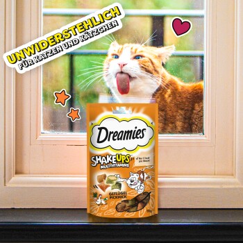 Emballage de friandises multivitaminées Dreamies Shake Ups pour chats avec un design ludique et le texte "Unwiderstehlich für Katzen und Kätzchen" près d'une fenêtre.