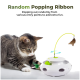 Getigerte Katze, die mit der Pfote ein weißes automatisches Katzenspielzeug mit grünem Ball und gelbem poppendem Bandfeder spielt; Text: 'Random Popping Ribbon'.
