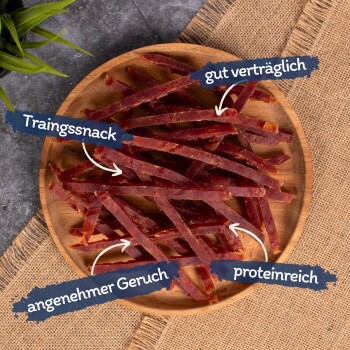 Trainingssnackstreifen, die auf einem Holzbrett angeordnet sind, mit Textetiketten, die "gut verträglich", "angenehmer Geruch" und "proteinreich" hervorheben.