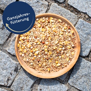 Eine Holzschüssel, gefüllt mit einer Vielzahl von Samen und Körnern, steht auf einer Steinoberfläche, mit einem runden Etikett, das "Ganzjahresfütterung" darüber liest.