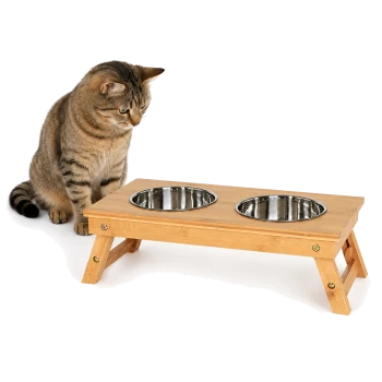 Getigerte Katze, die einen erhöhten Bambus-Futterspender mit zwei Edelstahlschalen vor weißem Hintergrund betrachtet.