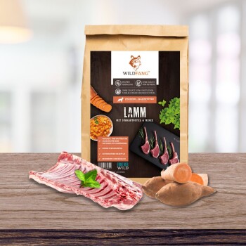 Verpackung von Wildfang Hundefutter mit Lamm, Süßkartoffeln und Minze, neben rohem Fleisch und Gemüse auf einer Holzoberfläche.
