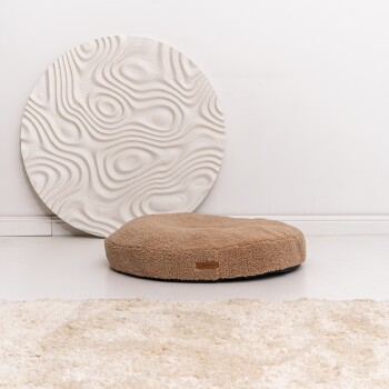 Ein runder, plüschiger Hundebett in hellbrauner Farbe steht auf einem weißen Boden, neben einer strukturierten weißen Wanddekoration.