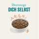 Schüssel mit Haustierfutter, gefüllt mit kleinen braunen Kroketten, mit dem Text 'Überzeuge DICH SELBST' darüber in fetter, verspielter Schrift.