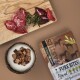 Ein Holzschneidebrett hält rohe Fleischstücke, frischen Rosmarin, eine Schüssel mit Haustierleckerlis und ein Paket mit der Aufschrift 'FALY PETFOOD TASTIES.'