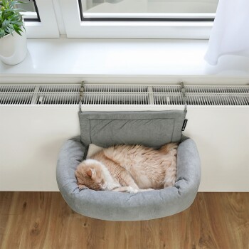 Eine flauschige, ingwerfarbene Katze, die friedlich in einem weichen grauen Haustierbett schläft, das an einer Fensterbank über einem Heizkörper befestigt ist, mit einer Topfpflanze in der Nähe.
