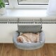 Eine flauschige, ingwerfarbene Katze, die friedlich in einem weichen grauen Haustierbett schläft, das an einer Fensterbank über einem Heizkörper befestigt ist, mit einer Topfpflanze in der Nähe.