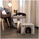 Eine dreifarbige Katze entspannt sich auf einem grauen Katzenbaum mit einem Kratzbaum und einem gemütlichen Versteck, neben einem modernen Stuhl und einer Lampe.