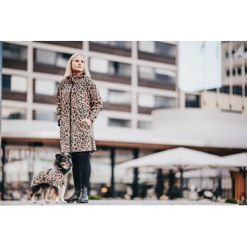 Eine Frau in einem Leopardenmuster-Mantel steht neben einem kleinen Hund, der eine passende Leopardenmuster-Jacke trägt, vor einem modernen urbanen Hintergrund.