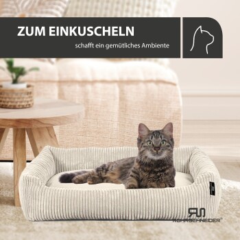Eine gemütliche Katze entspannt sich in einem gerippten Haustierbett mit einem cremefarbenen Kissen, neben einem Holztisch und einer Pflanze. Text: "ZUM EINKUSCHELN."