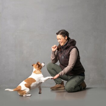 Eine Frau kniet auf einem grauen Boden und lächelt einen kleinen braun-weißen Hund an, der seine Pfote hebt. Sie trägt eine dunkle Weste und grüne Hosen.