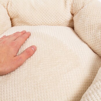 Eine Hand drückt sanft auf ein weiches, cremefarbenes Haustierbett mit einer strukturierten Oberfläche, das seinen plüschigen Komfort zeigt.