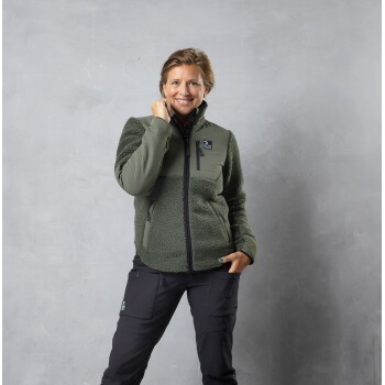 Eine Person, die eine grüne Fleecejacke mit Reißverschluss und schwarze Hosen trägt, posiert vor einem grauen Hintergrund.