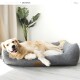 Ein Golden Retriever, der bequem auf einem grauen, gesteppten Haustierbett mit einem weißen Kissen liegt, umgeben von einer grünen Pflanze in einem hellen Raum.