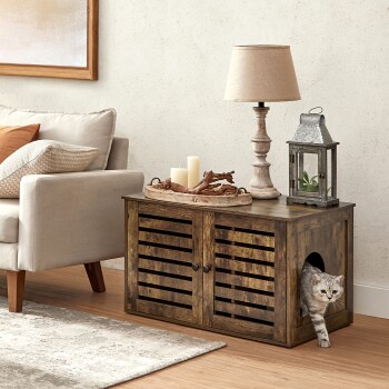 Eine graue getigerte Katze kommt aus einem Holzhaustierhaus mit lamellierten Türen, das neben einem beigen Sofa und dekorativen Gegenständen wie einer Lampe und einer Laterne steht.