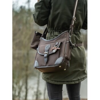 Eine Person in einer grünen Jacke trägt eine braune Haustiertragetasche mit einer vorderen Klappe, Seitentaschen und einem dekorativen Schlüsselanhänger.