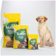 Drei gelb-grüne VUTTER!-Hundefuttertüten mit der Aufschrift 'PFLANZLICH VOLLWERTIG LECKER' und 'WIE HUHN' neben einem sitzenden Labrador.