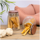 Ein Glasgefäß gefüllt mit Hundeleckerlis, einige fallen auf einen Holztisch, neben zwei weißen Seilspielzeugen und einem rosa Haustierbett im Hintergrund.