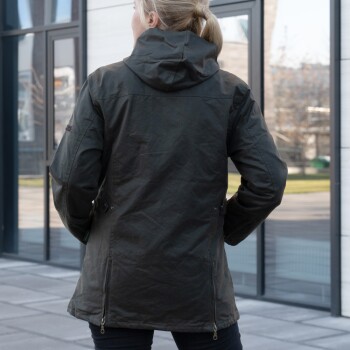 Frau in dunkelgrüner Kapuzenjacke steht im Freien mit Händen in den Taschen, Rückansicht, kein sichtbarer Text.