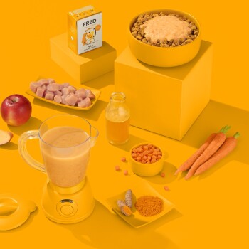 Ein gelbes Setup mit einer Schüssel Haustierfutter, die mit Sahne bedeckt ist, einem "FRED"-Kartons, frischem Hähnchen, Apfel, Karotten, Gewürzen, einem Mixer und einer Saftflasche.