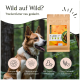 Ein glücklicher Hund sitzt neben einer bunten "Wild" Haustierfutterverpackung mit Hirschillustrationen und Etiketten für hypoallergen, getreidefrei und von Experten entwickelt.