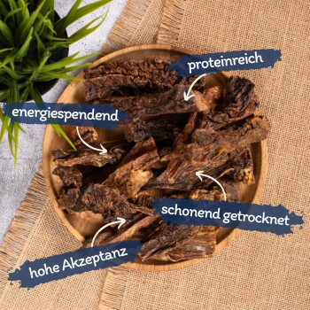 Eine Holzschüssel mit getrockneten Fleischstreifen, beschriftet auf Deutsch, mit einer grünen Pflanze im Hintergrund. Die Etiketten beinhalten "reich an Protein" und "energiegebend."