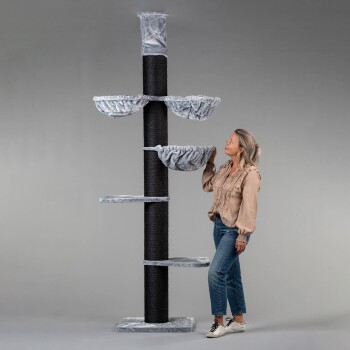 Ein hoher, mehrstöckiger Kratzbaum mit weichen, grau bezogenen Plattformen und einem zentralen schwarzen Kratzpfosten, neben einer Frau, die ihn bewundert.