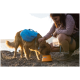Eine Person kniet neben einem Hund mit einem blauen Rucksack und füttert ihn aus einem orangen Napf mit der Aufschrift "JAFWREAR" in der Nähe eines Gewässers bei Sonnenuntergang.