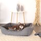 Ein entspannter brauner Chihuahua liegt in einem gemütlichen grauen Haustierbett auf einem weichen Teppich, mit einem modernen Tisch und dekorativen Vasen im Hintergrund.