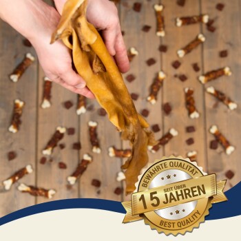 Eine Hand hält einen kaubaren Hundesnack zwischen kleinen knochenförmigen Snacks auf Holz, mit einem Abzeichen, das "Beste Qualität seit über 15 Jahren" sagt.