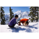 Eine Person in einem lila Shirt kniet im Schnee und füttert einen Hund in einer orangefarbenen Jacke, mit schneebedeckten Bergen im Hintergrund.