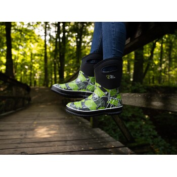 Ein Paar grün und schwarz gemusterte Gummistiefel, die auf einer Holzbrücke ruhen, mit einem verschwommenen Wald im Hintergrund.