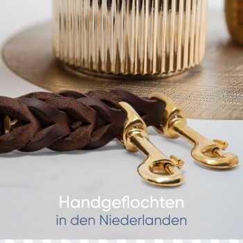 Eine geflochtene braune Lederleine für Hunde mit goldenen Verschlüssen, die auf einer strukturierten goldenen Oberfläche liegt, mit dem Text "Handgeflochten in den Niederlanden" darunter.
