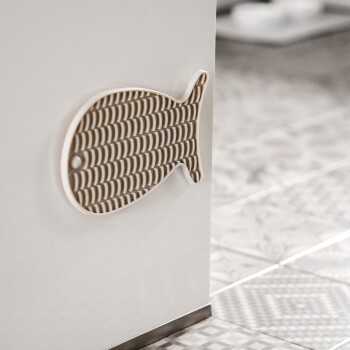 Ein dekorativer wandmontierter Fischhaken in Beige und Weiß, der an einem weißen Schrank angebracht ist, mit gemustertem Boden im Hintergrund.