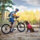 Ein Radfahrer in einem grauen und orangefarbenen Shirt steht neben einem Mountainbike, mit einem braunen Hund in einem roten Rucksack, der neben ihm im Wald sitzt.
