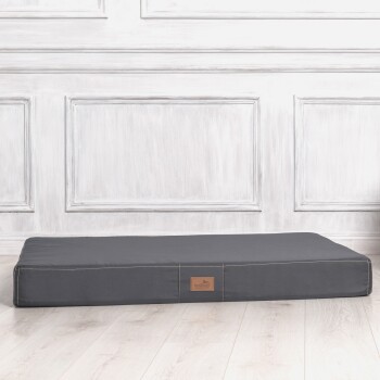 Ein dunkelgraues Haustierbett mit einer gesteppten Kante und einem kleinen Lederetikett mit einem Hundelogo, vor einem hellen Holzboden und einer weißen Paneelwand.