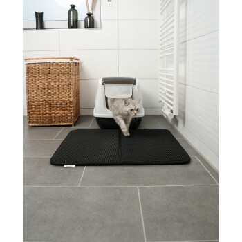 Eine graue Katze kommt aus einer weißen Katzentoilette auf eine schwarze Matte, mit einer natürlichen Holzdekoration im Hintergrund.