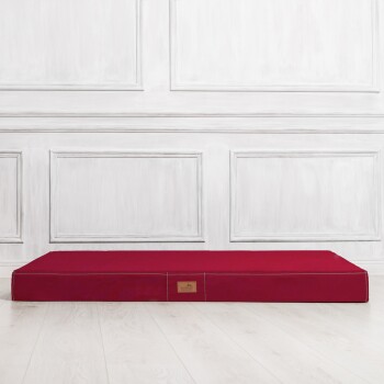 Rotes rechteckiges Tierbett mit genähtem Rand und einem kleinen Logo-Anhänger, auf einem hellen Holzboden vor einer weißen Paneelwand platziert.