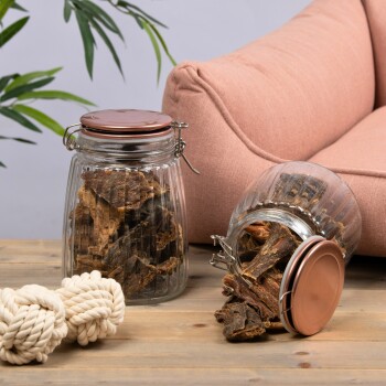 Zwei Glasbehälter gefüllt mit Hundeleckerlis, einer umgekippt, neben einem weichen rosa Haustiersofa und zwei weißen Seilspielzeugen auf einer Holzoberfläche.