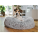 Kleiner dreifarbiger Hund mit unscharfem Gesicht sitzt in einem runden, flauschigen grauen Hundebett auf einem Holzboden in einem hellen Wohnzimmer.