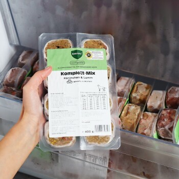 Hand hält eine gefrorene Hundefutterpackung mit der Aufschrift 'Komplett‑Mix Kaninchen & Lamm' (BARFER'S) 400 g im Gefrierschrank.