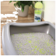 A gray cat litter box filled with gray litter and green clumping agents, with a plant and a bottle of cleaning solution in the background.