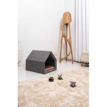 Ein gemütliches graues Haustierhaus mit einem weichen rosa Kissen innen, begleitet von drei grauen Filzspielzeugen, auf einem plüschigen beigen Teppich platziert.