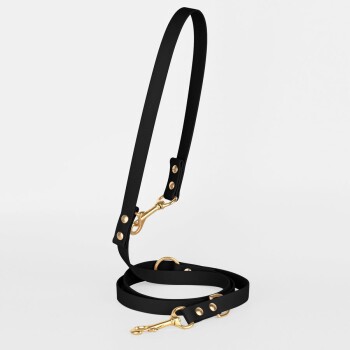 Eine Person in einem dunklen Mantel und hohen schwarzen Stiefeln steht auf einer Kopfsteinpflasterstraße und hält eine Leine, die an einem sitzenden braunen Hund befestigt ist.